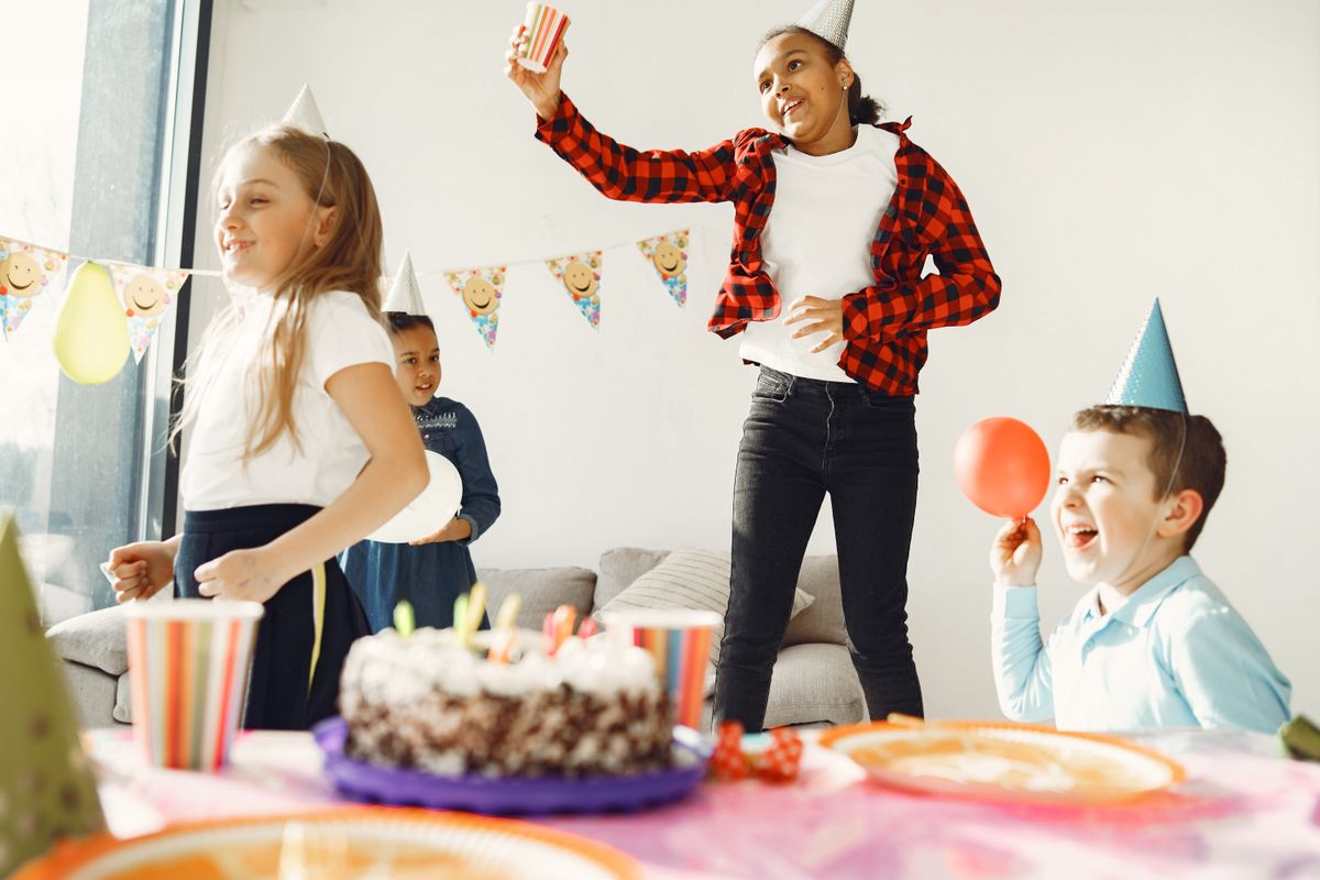 Children in party hats dancing around a birthday cake at a party at home