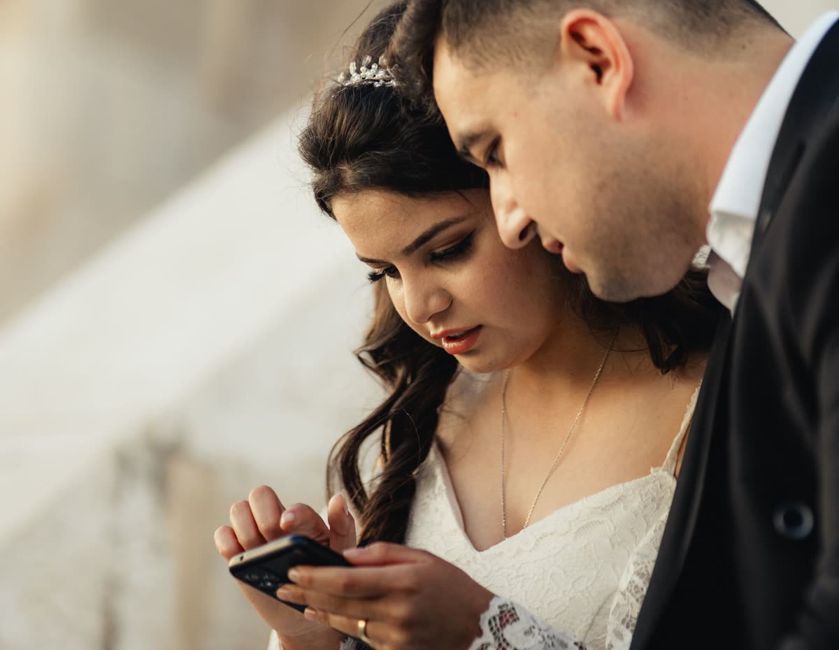 A bride and groom in wedding attire leaning together to look at photos on a phone