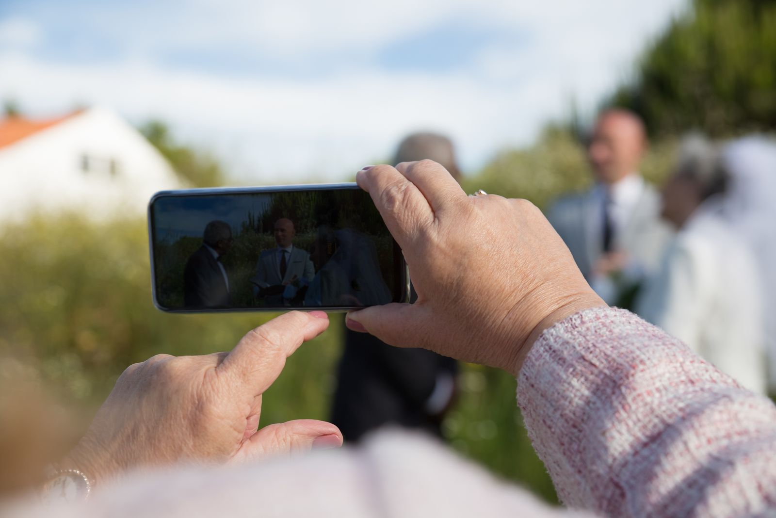 An older wedding guest’s hands holding a phone up to film the couple during an outdoor ceremony