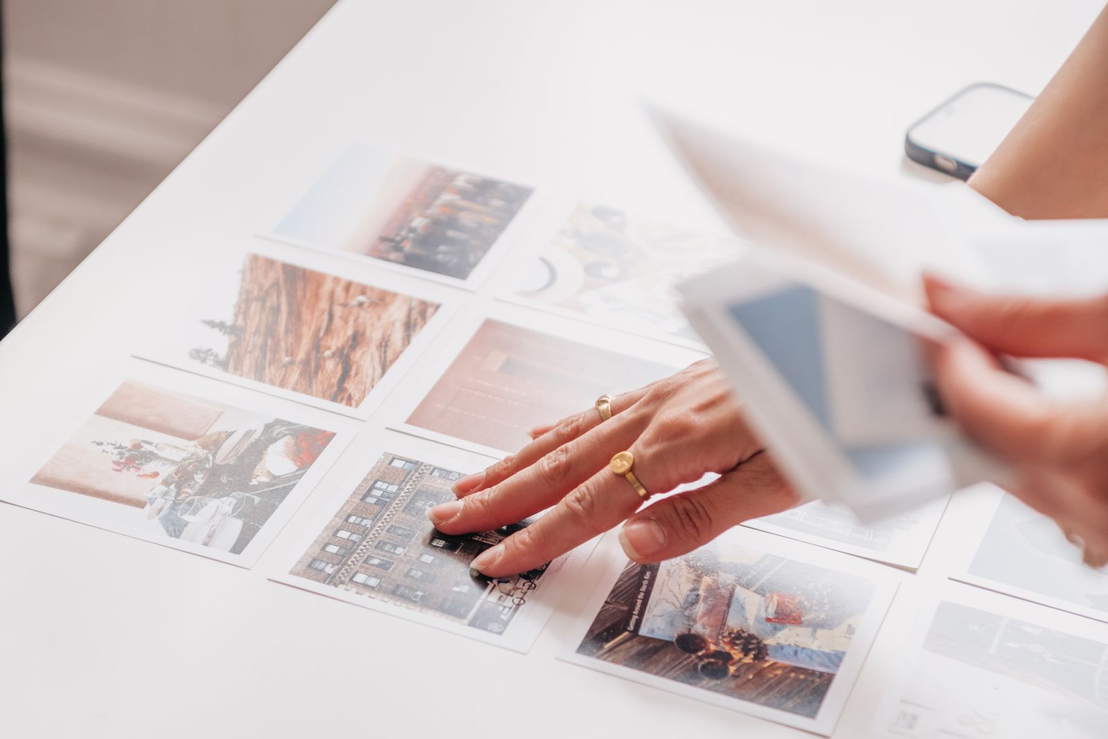 Hands sorting a grid of printed photographs on a white table, a phone resting at the edge