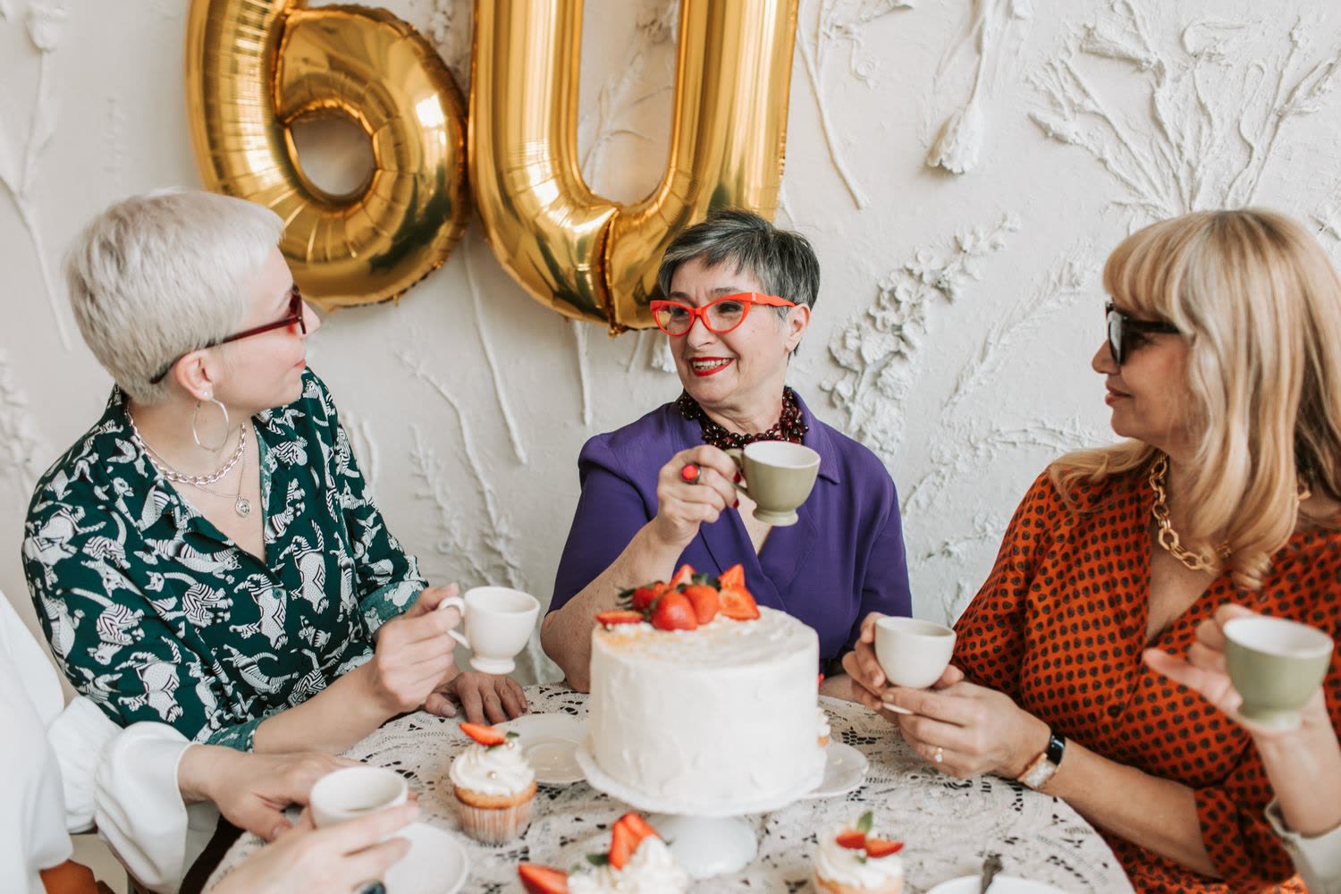Three friends celebrating a 60th birthday with cake and tea under gold number balloons