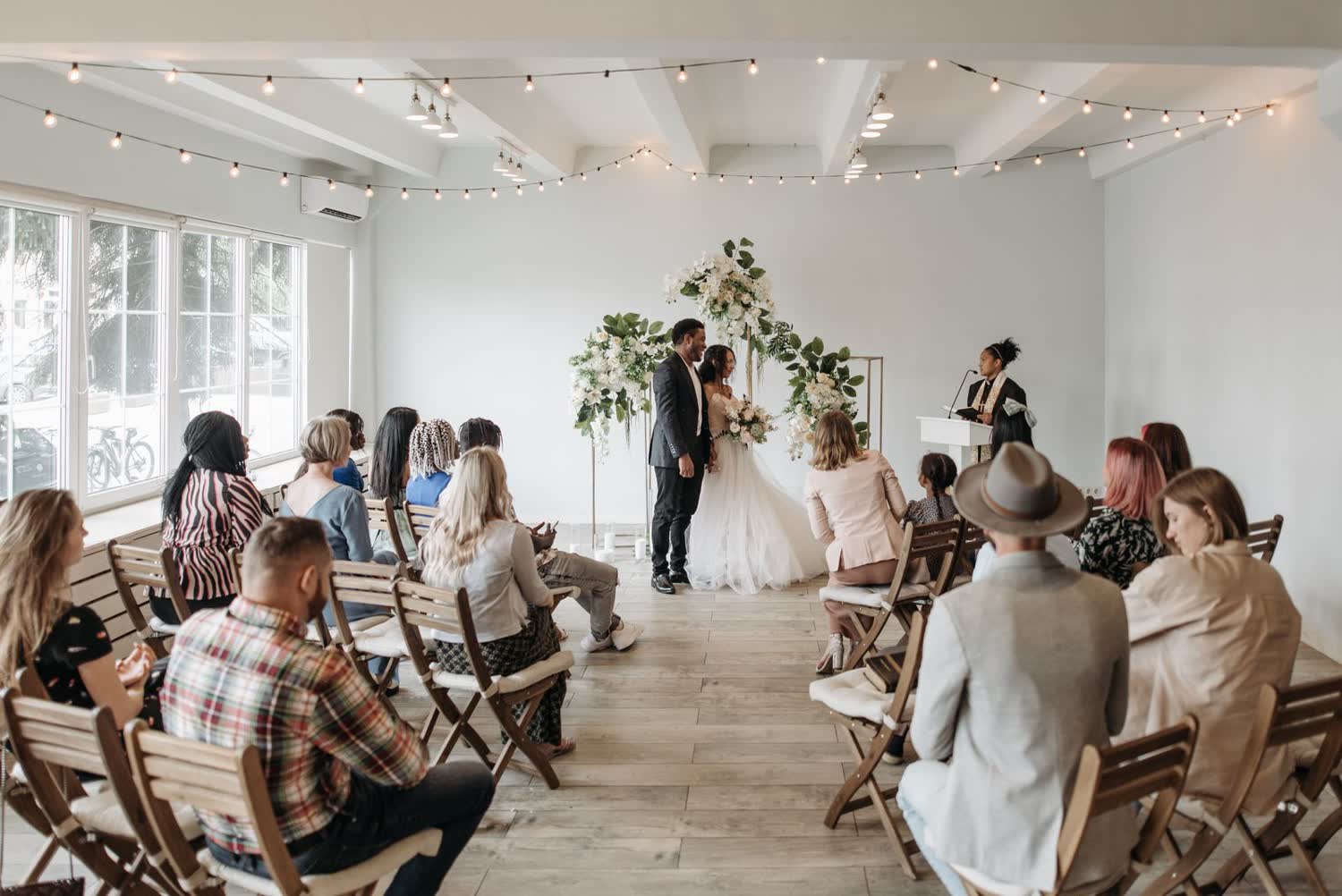 Wedding guests seated on wooden chairs at an indoor ceremony, watching the couple rather than holding up phones