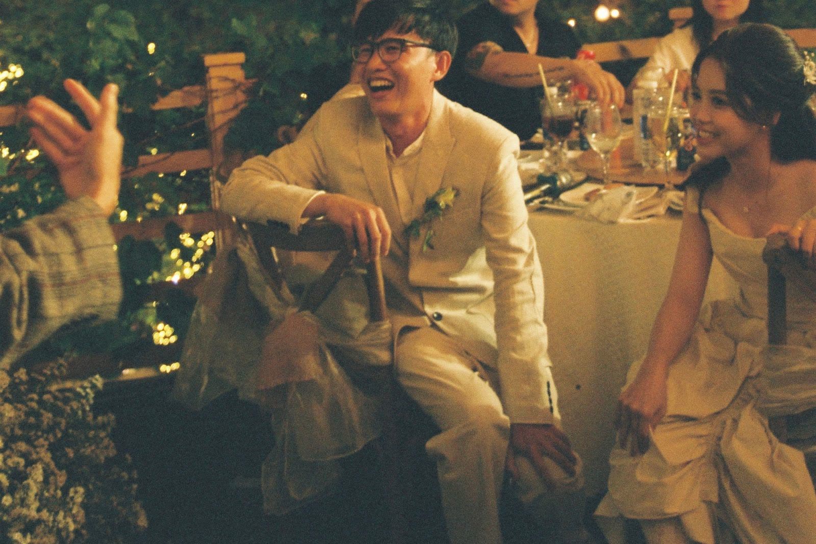 A couple sitting at their reception table at night under string lights, the groom laughing as a guest gestures towards them