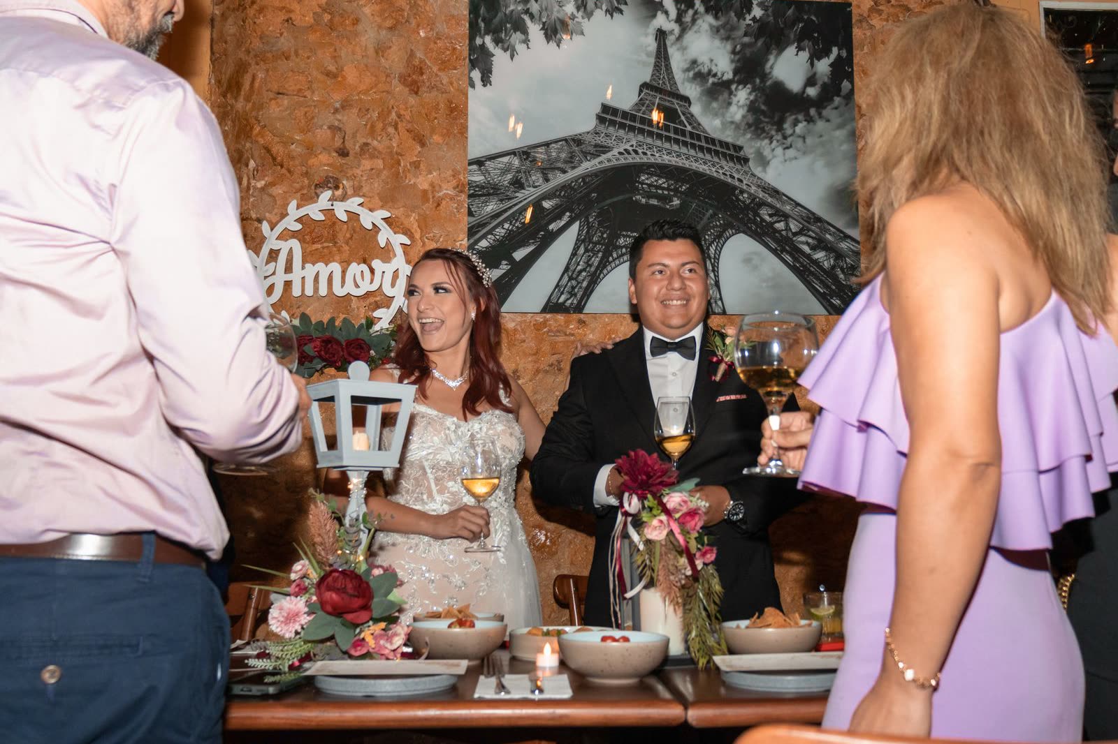 A bride and groom laughing at a small restaurant table while a handful of guests raise their glasses around them