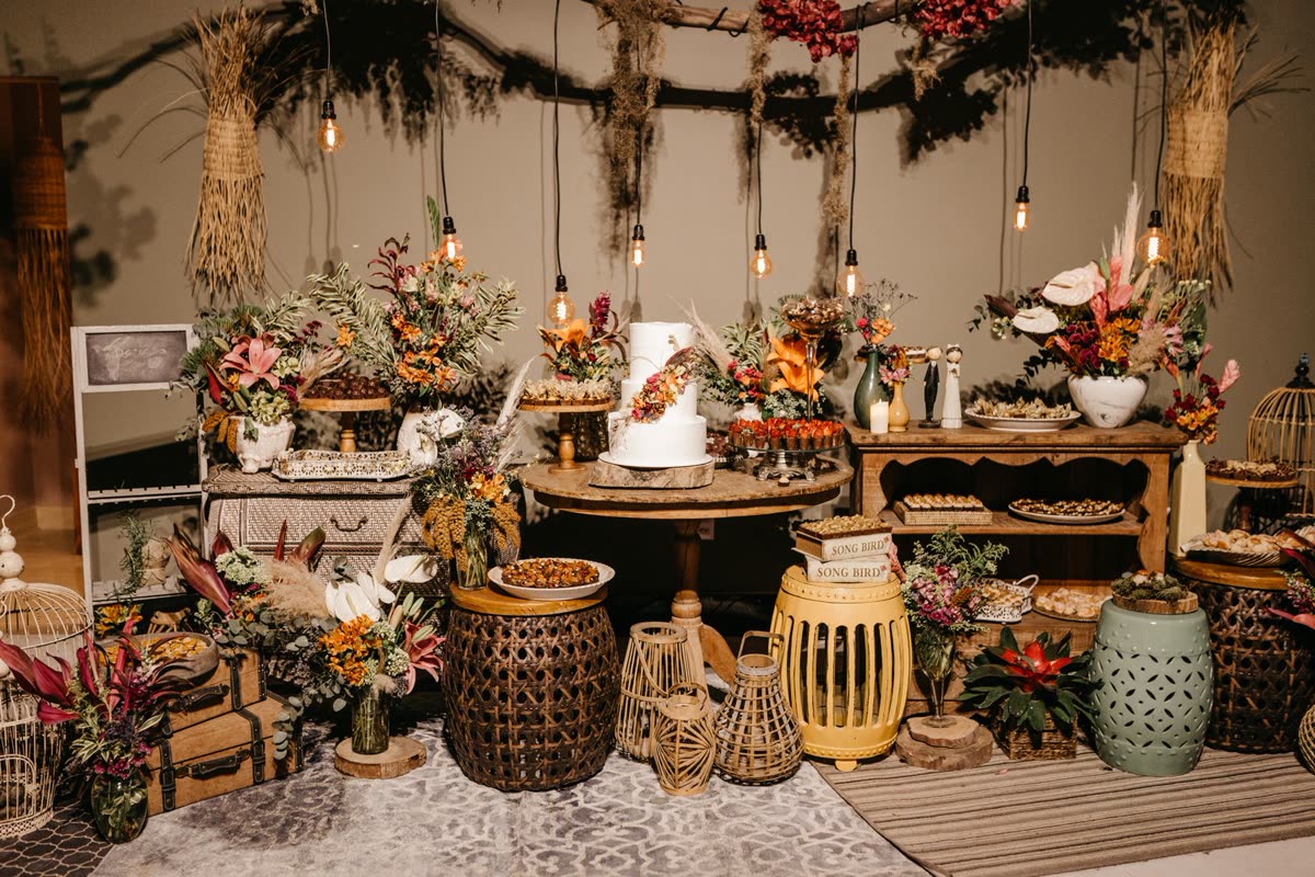 A rustic wedding dessert table with a white cake, dried flowers and lanterns under hanging bulbs