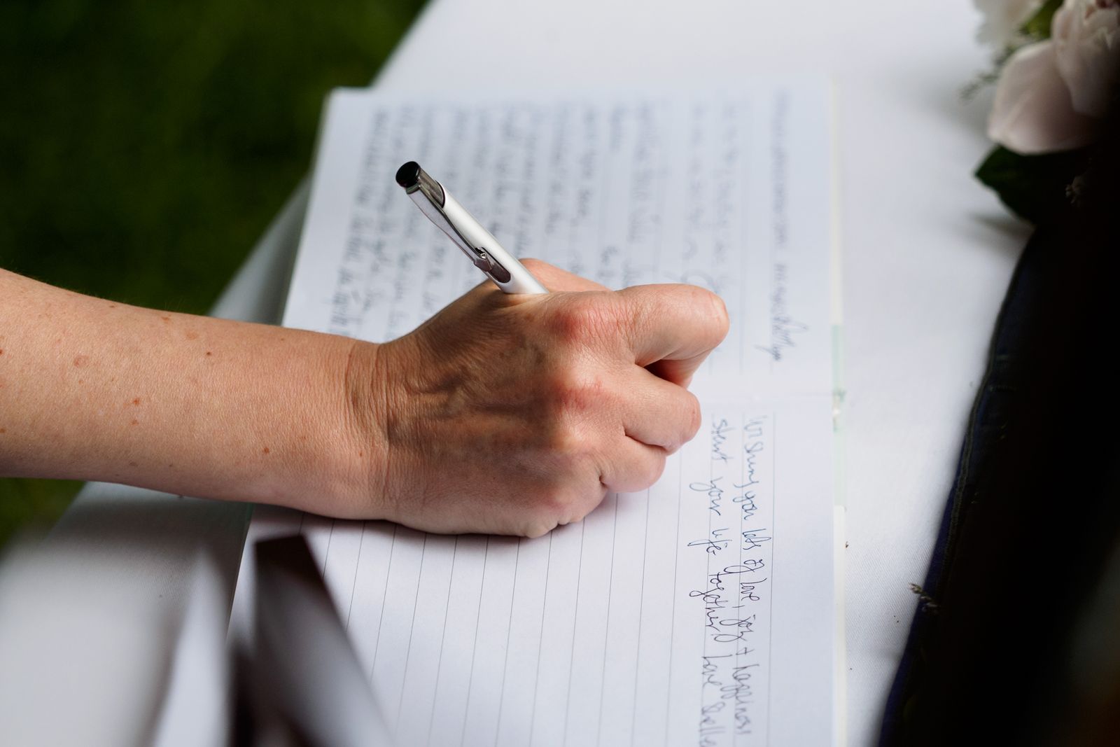 A hand writing a heartfelt message in a wedding guest book with a pen, a bouquet visible nearby