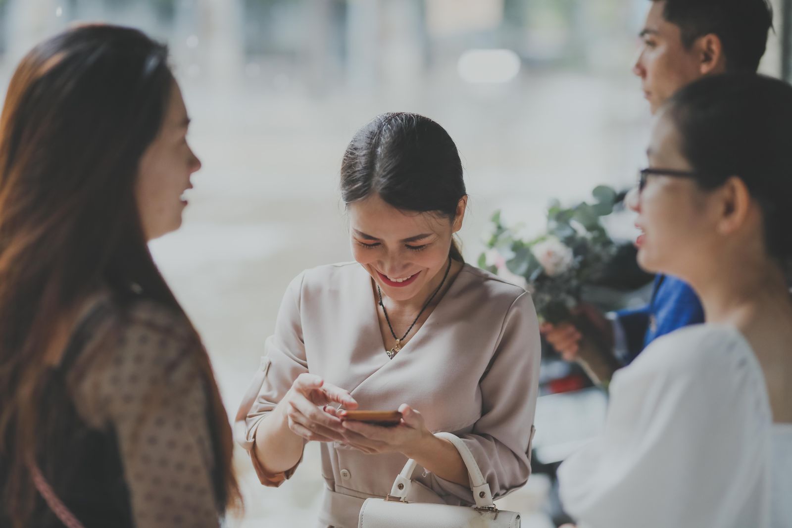 A wedding guest smiling down at her phone while standing with other guests, a bouquet visible behind her