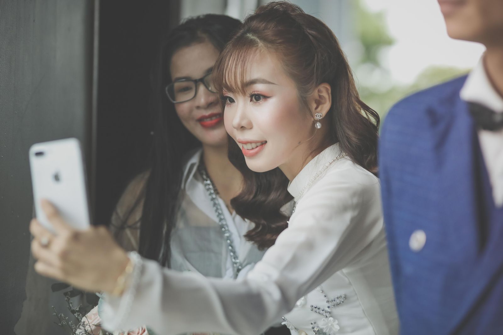 A bride in white and a wedding guest leaning in together to capture a moment on a phone
