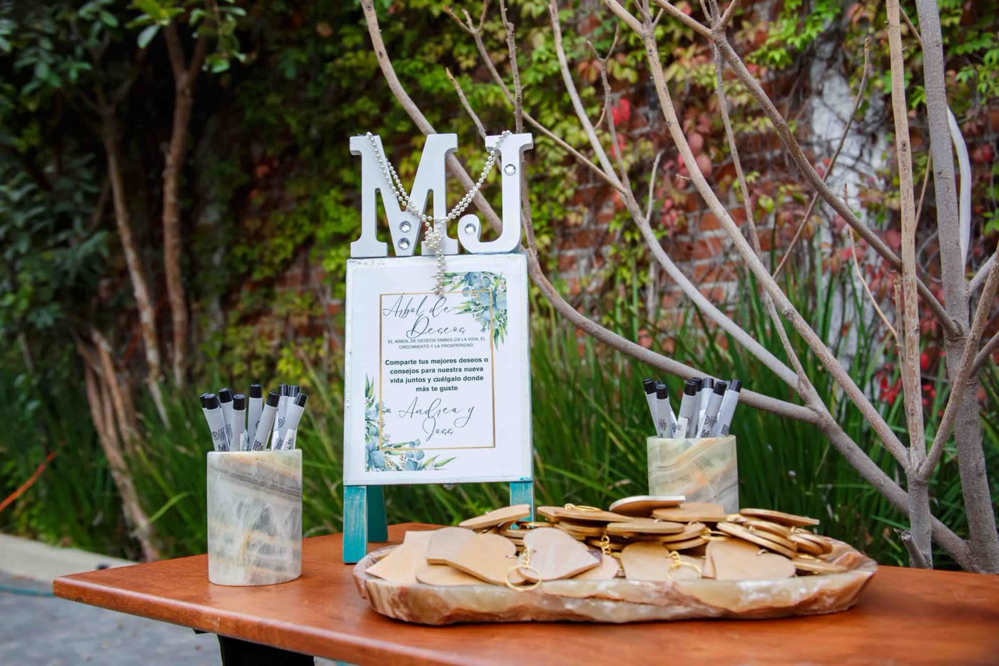 A wedding message station on a wooden table with a decorative sign, pots of pens and a tray of wooden heart tags for guests to write their wishes on