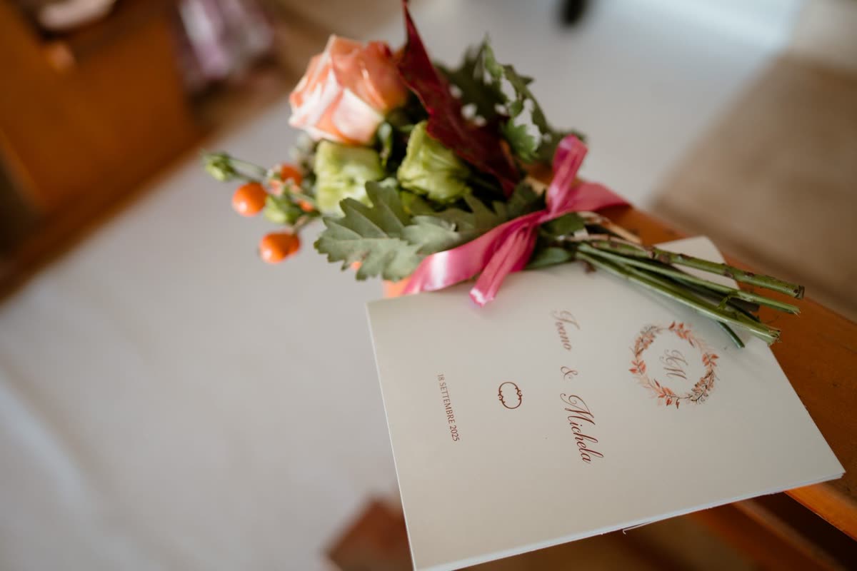 A cream printed order of service resting on a church pew beside a small posy of peach roses tied with pink ribbon