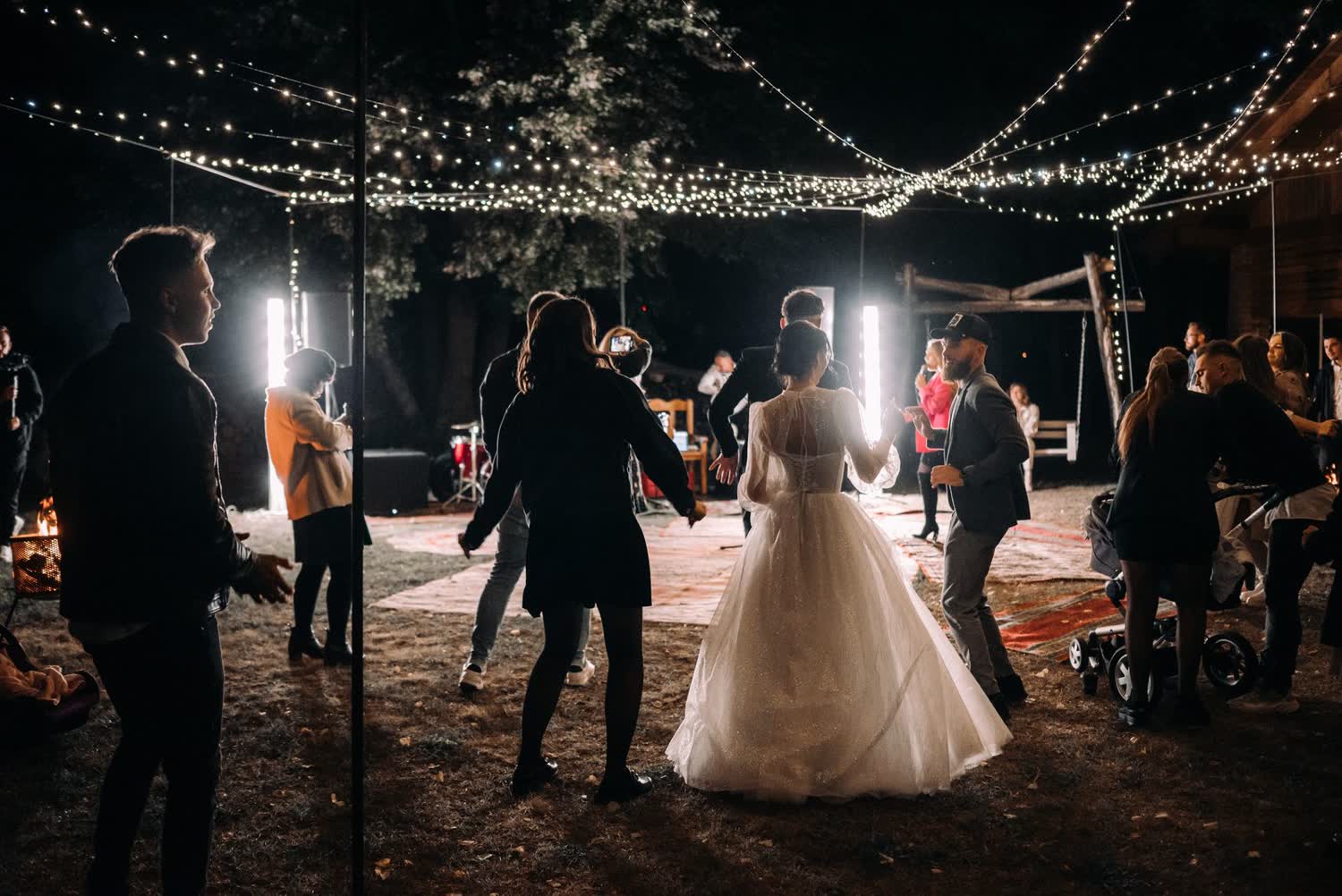 Wedding guests dancing outdoors at night under string lights, with one guest filming on a phone