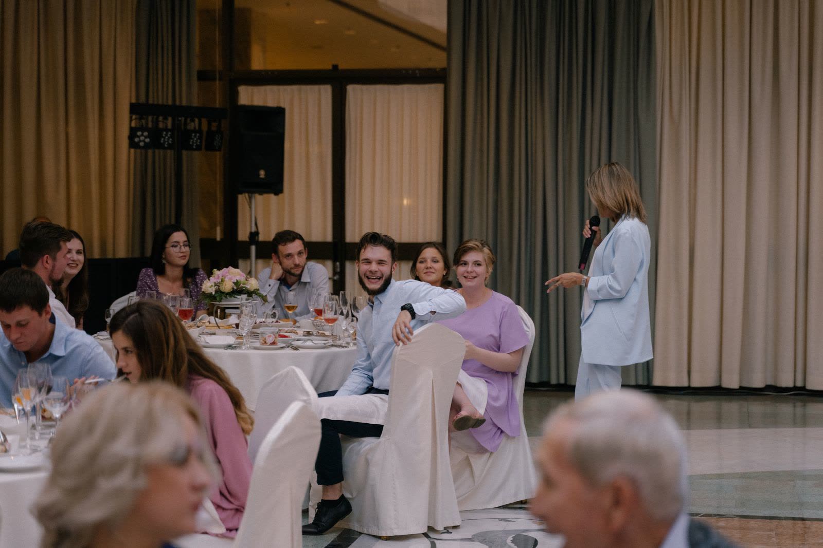 A host with a microphone reading out to a room of wedding guests at round tables, one guest turned round in his chair laughing