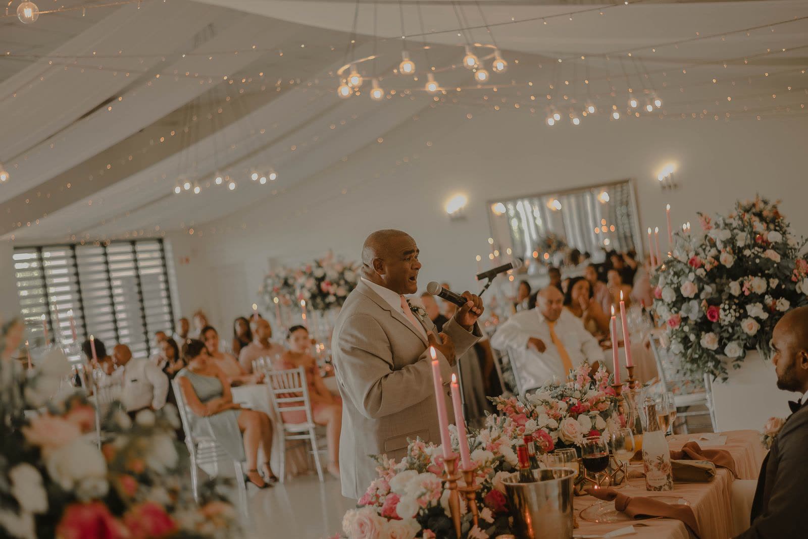 A guest in a beige suit giving a toast with a microphone at a wedding reception under string lights, guests watching from tables