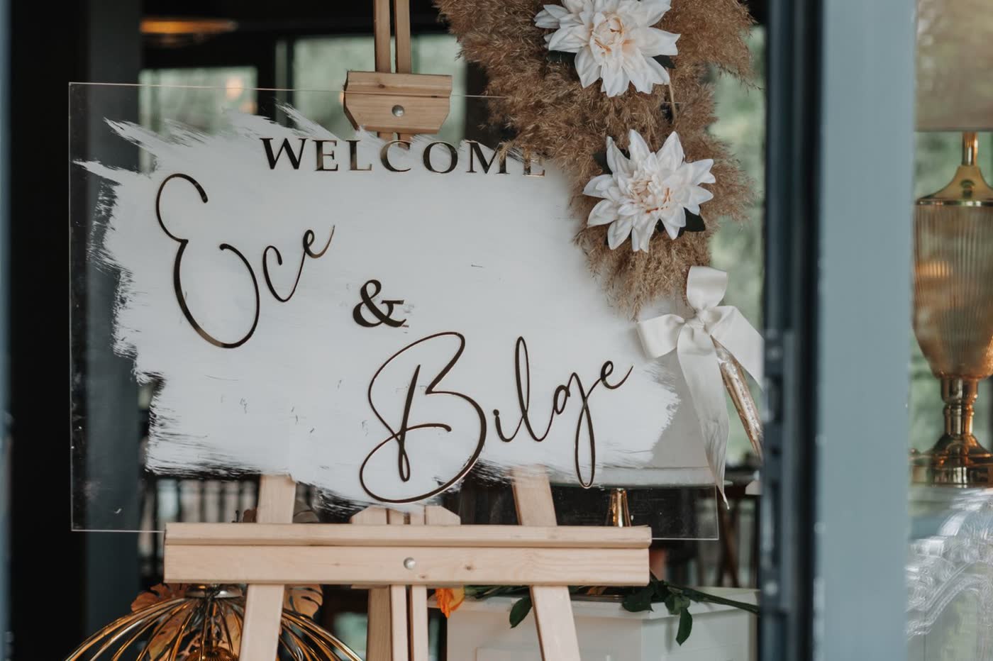 A wedding welcome sign propped on a wooden easel at a venue entrance, decorated with pampas grass and white flowers